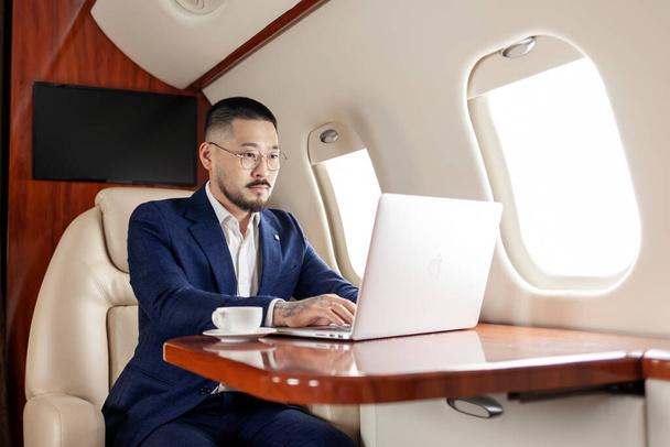 successful Asian businessman in suit and glasses flies in private luxury plane with laptop, Korean trader financier works at computer in airplane, luxury lifestyle - Fotografie, Obrázek