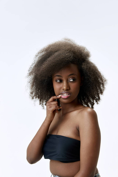 Portret van een mooie, jonge, Afrikaanse vrouw met krullend haar, goed onderhouden, gladde, gezonde huid die poseert tegen een witte studioachtergrond. Begrip natuurlijke schoonheid, huidverzorging, gezondheid, mode, wellness, reclame - Foto, afbeelding