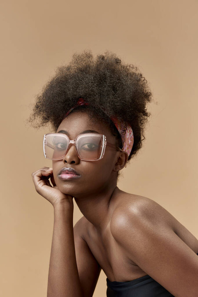 Πορτρέτο της νεαρής, κομψής, όμορφης, αφρικανικής γυναίκας στην κορυφή και μοντέρνα γυαλιά ηλίου με φόντο στούντιο. Έννοια της φυσικής ομορφιάς, φροντίδα του δέρματος, υγεία, μόδα, ευεξία, διαφήμιση - Φωτογραφία, εικόνα