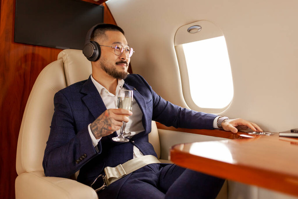 επιτυχημένος Ασιάτης επιχειρηματίας πετά σε ιδιωτικό τζετ πολυτελείας με ένα ποτήρι σαμπάνια και ακούει μουσική στα ακουστικά, Κορεάτης επιχειρηματίας με κοστούμι ξεκουράζεται και χαλαρώνει στο αεροπλάνο, πολυτελή τρόπο ζωής - Φωτογραφία, εικόνα