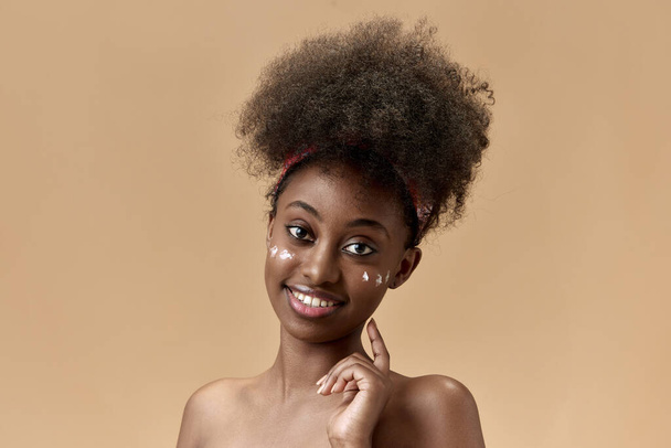 A Spf le importa. Retrato de mujer joven, sonriente, hermosa, africana que aplica crema hidratante facial sobre el fondo del estudio. Concepto de belleza natural, cuidado de la piel, salud, moda, bienestar, anuncio - Foto, Imagen