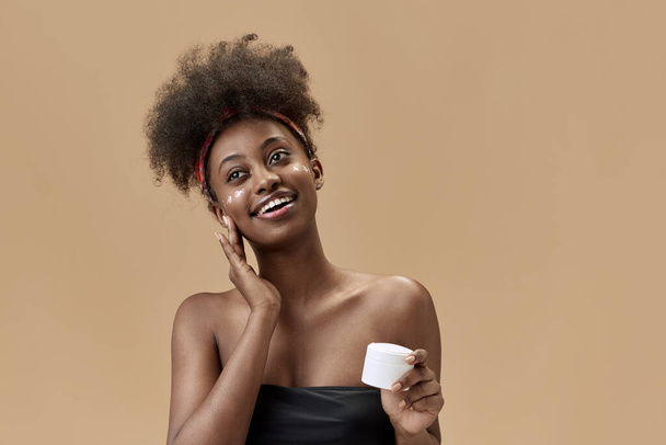 Πορτρέτο της νεαρής, θετικής, όμορφης, αφρικανικής γυναίκας που εφαρμόζει ενυδατική κρέμα προσώπου σε φόντο στούντιο. Έννοια της φυσικής ομορφιάς, φροντίδα του δέρματος, υγεία, μόδα, ευεξία, διαφήμιση - Φωτογραφία, εικόνα