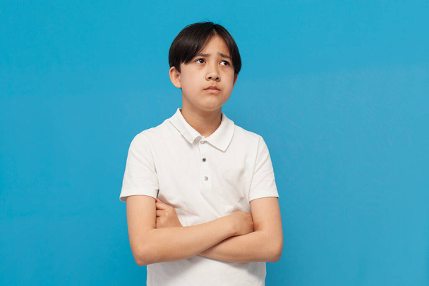 δυστυχισμένο αγόρι από την Ασία των δώδεκα ετών είναι λυπημένο και προσβεβλημένο σε μπλε απομονωμένο φόντο, δυσαρεστημένο κορεάτικο παιδί με λευκό μπλουζάκι στέκεται με σταυρωμένα τα χέρια και σκέφτεται - Φωτογραφία, εικόνα