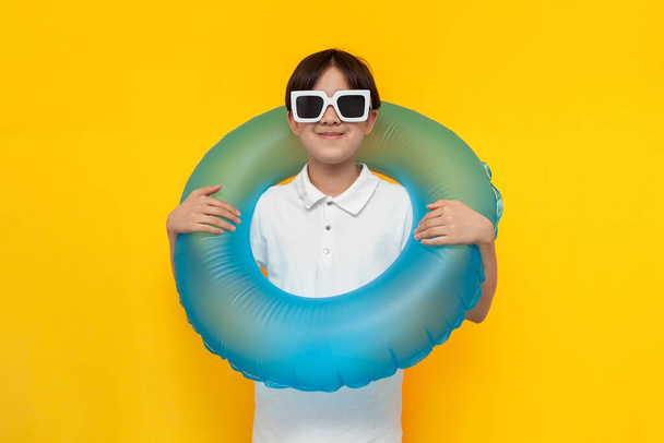 tiener jongen van twaalf jaar oud in zonnebril houdt opblaasbare zwemring op gele geïsoleerde achtergrond, kind in de zomer op het strand, het concept van reizen naar de zee en zwemmen - Foto, afbeelding