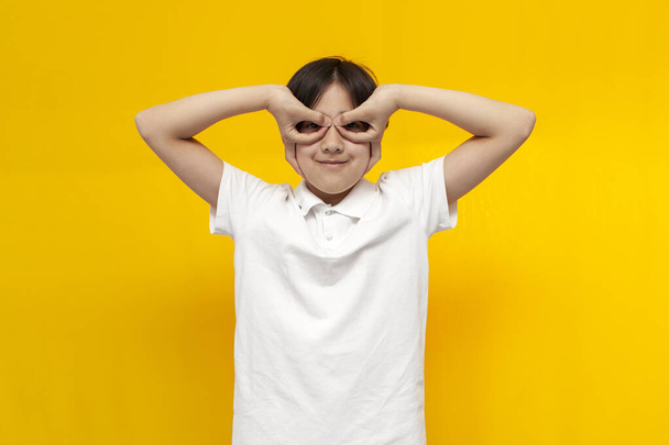 asian boy of twelve years old in white t-shirt makes mask over his eyes with his hands and fools around on yellow isolated background, korean child plays and looks through glasses from his fingers - Фото, изображение