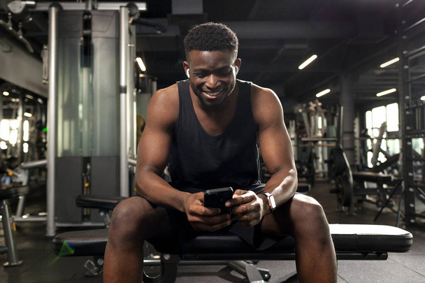 mladý atletický africký Američan sedí v tmavé tělocvičně a používá smartphone, sportovní týpci typy on-line na telefonu ve fitness klubu a usmívá - Fotografie, Obrázek