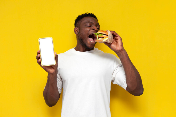 Afričan Američan v bílém tričku drží velký burger a ukazuje prázdné obrazovky smartphone na žlutém izolovaném pozadí, mladý člověk jí rychlé občerstvení a inzeruje telefonní displej, kopírovací prostor - Fotografie, Obrázek