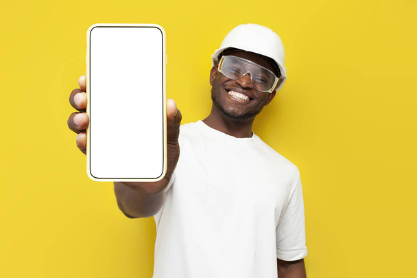 männlich afrikanisch-amerikanischer Bauunternehmer in Uniform zeigt leeren Smartphone-Bildschirm auf gelbem Hintergrund, Arbeiter Ingenieur Architekt mit Hut und Schutzbrille wirbt mit Kopierraum am Telefon, Attrappe - Foto, Bild