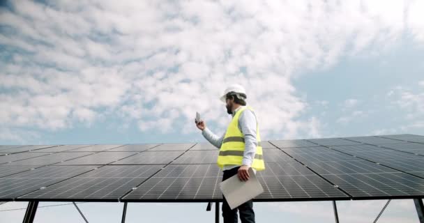 Mężczyzna inżynier mówi na smartfonie na farmie słonecznej. Niskokątny mężczyzna w mundurze spacerujący w pobliżu paneli fotowoltaicznych i odbierający telefon przed zachmurzonym błękitnym niebem w słoneczny dzień na słonecznej elektrowni. - Materiał filmowy, wideo