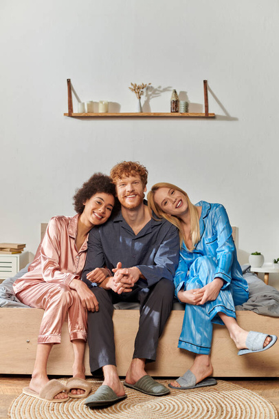 open relationship concept, polygamy, understanding, three adults, happy man and multicultural women in pajamas sitting on bed at home, cultural diversity, acceptance, bisexual  - 写真・画像