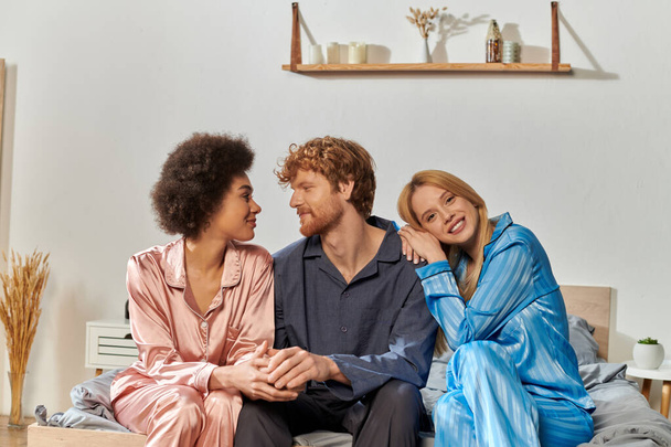 polygamy concept, understanding, three adults, redhead man and multicultural women in pajamas sitting on bed at home, cultural diversity, acceptance, bisexual, positive, open relationship  - Photo, Image