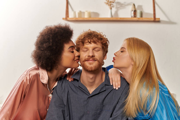 polygamy concept, open relationship, portrait of three adults, multicultural women kissing happy redhead man, polyamorous family in pajamas, cultural diversity, acceptance, bisexual, polyamory  - Фото, изображение