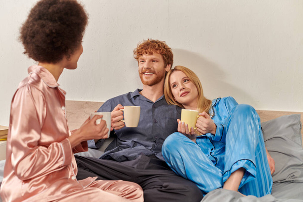 concepto de relación abierta, pelirroja y mujeres interracial en pijama sosteniendo tazas de café de la mañana, estilo de vida, bisexual, comprensión, tres adultos, diversidad cultural, aceptación  - Foto, Imagen