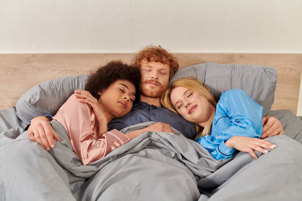 open relationship, polygamy, understanding, three adults sleeping under blanket, redhead man and multicultural women in pajamas, lovers, morning, cultural diversity, acceptance, bisexual  - 写真・画像