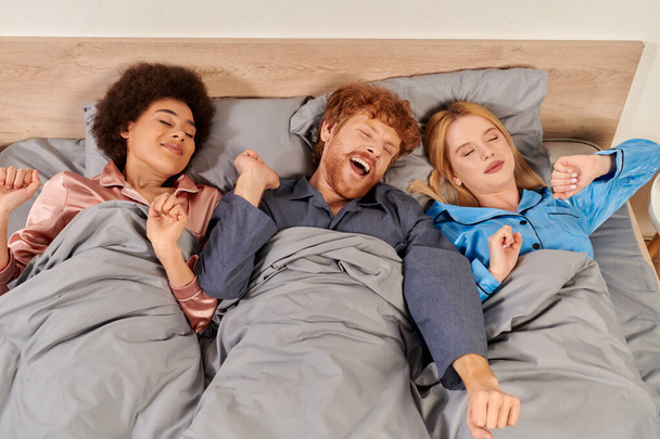 полиаморная концепция, три взрослых, мужчина и межрасовые женщины в пижамах просыпаются вместе, утром, под одеялом, спальня, культурное разнообразие, бисексуалы, открытые отношения, полигамия, вид сверху - Фото, изображение