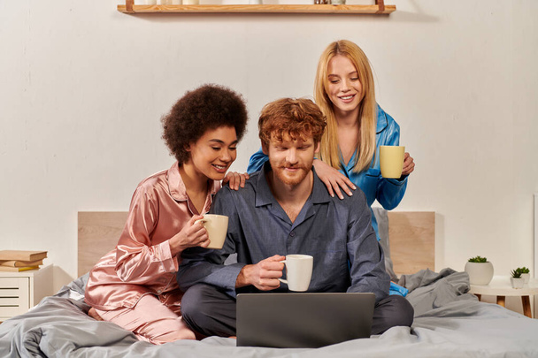 poligamia, mujeres multiculturales y pelirrojo hombre en pijama viendo la película en el ordenador portátil, la celebración de tazas de café en el dormitorio, diversidad cultural, aceptación, bisexual, relación abierta  - Foto, Imagen