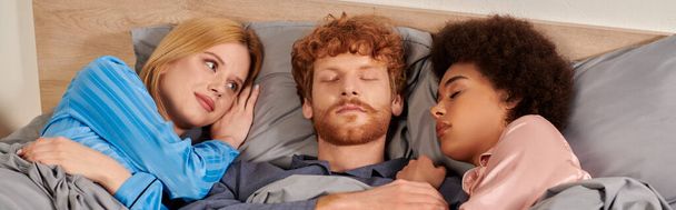 любовний трикутник, полігамія, три дорослі сплять разом, рудий чоловік і мультикультурні жінки в піжамі, спальні, культурне різноманіття, прийняття, бісексуали, банер - Фото, зображення