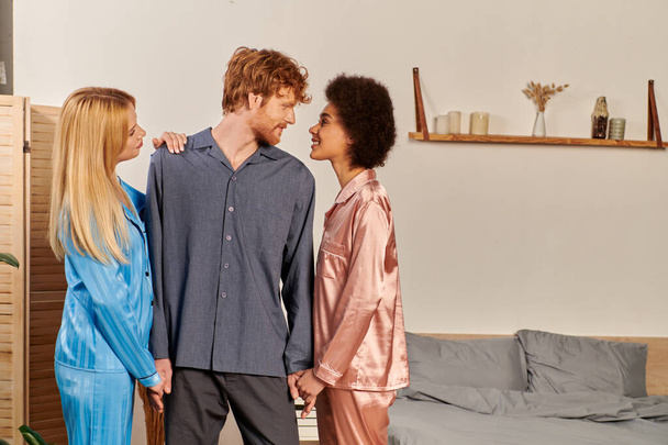 polygamy lovers in pajamas, happy man with red hair holding hands of interracial women at home, non traditional partners, freedom in relationship, acceptance and understanding  - Photo, Image
