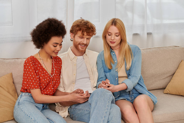modern family, polygamy concept, freedom in relationship, cultural diversity, redhead man holding hands with multicultural women, sitting on couch in living room, polyamorous lifestyle  - Photo, Image