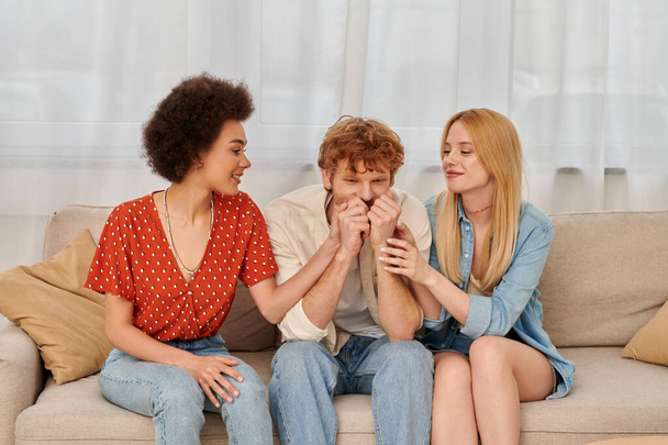 polyamorous relationship, cultural diversity, redhead man sitting on couch with happy multiracial female lovers, freedom and acceptance, love triangle, three people in open relationship  - Photo, Image