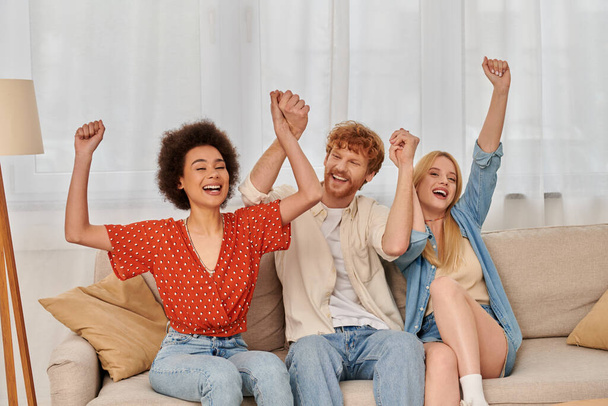 polyamorous relationship, cultural diversity, redhead man sitting on couch with happy multiracial female lovers, raised hands, freedom and acceptance, love triangle, people in open relationship  - 写真・画像