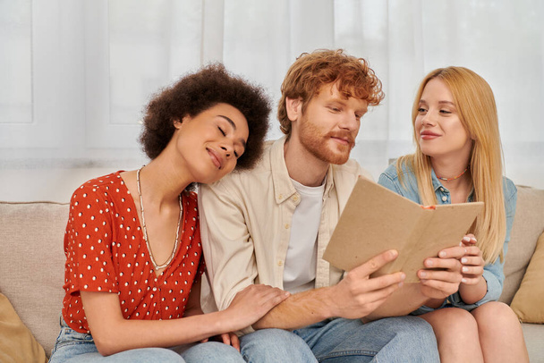 polygamy, open relationships, redhead man reading book to pleased multicultural women in living room, non traditional lovers, cultural diversity, romance and love, modern family concept  - Photo, Image