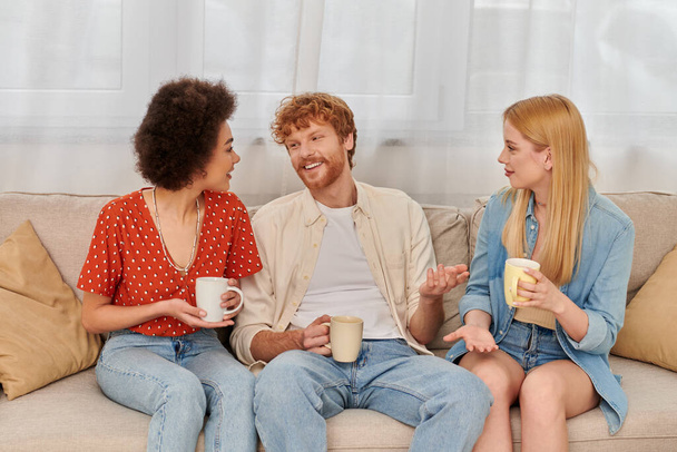 concepto poliamoroso, diversidad de relaciones, amantes de la poligamia felices sentados en el sofá y sosteniendo tazas de café, hombre y mujeres interracial en la sala de estar, bisexuales y personas poligamia  - Foto, imagen