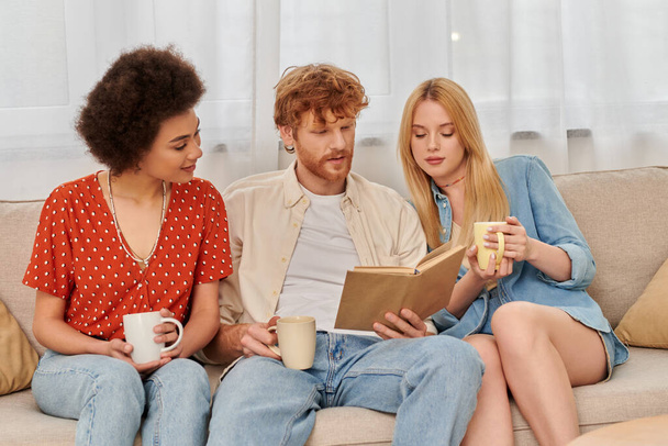 alternative relationships, polygamy, intelligent redhead man reading book to multicultural women in living room, modern family, hobby and leisure, polyamorous family, cups of coffee  - Photo, Image
