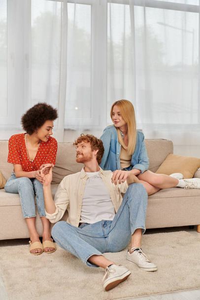 love triangle concept, diversity in relationships and culture, non monogamy, cheerful redhead man sitting near beautiful multicultural women in living room, lovers, modern family, open relationship  - Foto, afbeelding