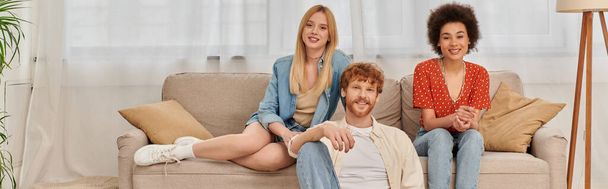 love triangle concept, diversity in relationships and culture, non monogamy, happy redhead man and multicultural women looking at camera in living room, lovers, acceptance, open relationship, banner  - Foto, Imagem