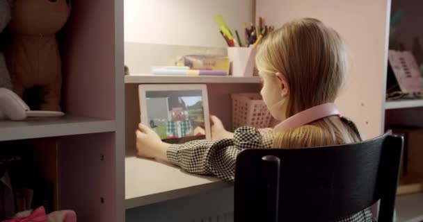 La niña de 7 años está jugando en la habitación de los niños con auriculares rosados y utilizando la tableta de computadora digital, disfrutando de contenido fresco de vídeo o foto en la red social, jugando juegos en línea. - Metraje, vídeo
