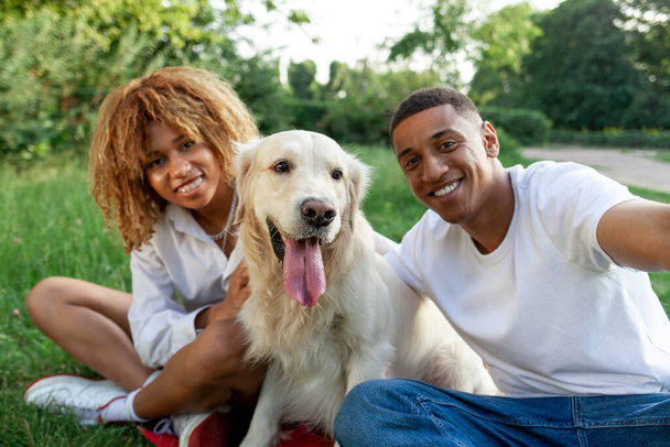 Afrikaans Amerikaans jong stel zitten in park samen met hond en selfie nemen, man en vrouw met golden retriever communiceren via video call in de natuur, gelukkig gezin met huisdier - Foto, afbeelding