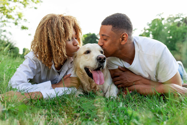 african american happy couple lie together with dog in park in summer, man and woman kiss and love golden retriever in nature, young family with pet outdoors - Фото, зображення