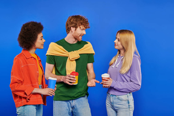 alternative relationships, polyamory lovers with coffee, young redhead man and beautiful multiracial women holding paper cups on blue background, studio shot, takeaway drink, polygamy people - Valokuva, kuva