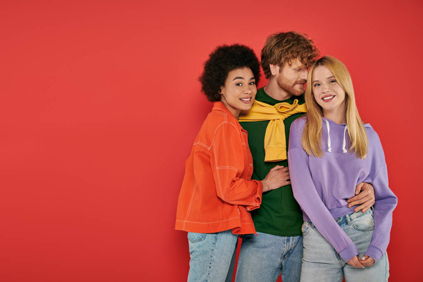 polyamorous relationship concept, polygamy lovers, young man and multicultural women hugging on coral background, studio shot, denim fashion, love triangle, bonding and acceptance  - Φωτογραφία, εικόνα