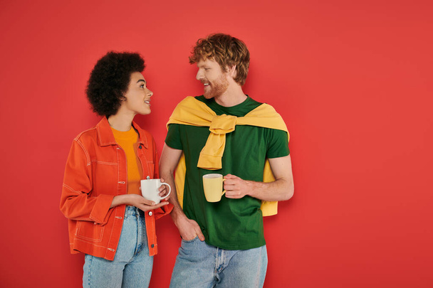 multicultural couple holding cups of coffee on coral background, morning routine, cultural diversity, vibrant colors, stylish outfits, interracial lovers holding mugs and having conversation  - Foto, afbeelding