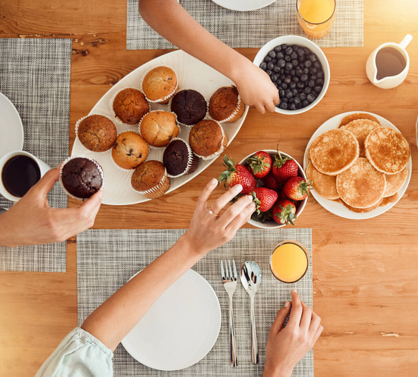 Breakfast, food and hungry people in dining room, eating healthy and above table setting or home in the morning. Fruit, pancakes and hands on strawberry or muffin plate for nutrition or diet. - Fotó, kép