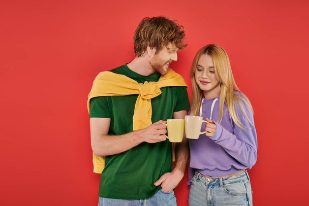 happy young couple holding cups of coffee on coral background, morning routine, vibrant colors, stylish outfits, modern family, people holding mugs with hot drink, husband and wife  - Photo, Image