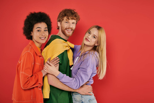 open relationship, polygamy concept, three interracial lovers hugging each other on coral background, cultural diversity, polyamorous, happy multiethnic people looking at camera  - Foto, Bild