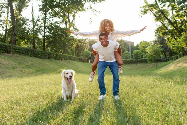 Afrikaans Amerikaans jong paar verliefd loopt met hond in het park in de zomer en glimlacht, de man draagt zijn vriendin op zijn rug en loopt met golden retriever buiten op het gras - Foto, afbeelding