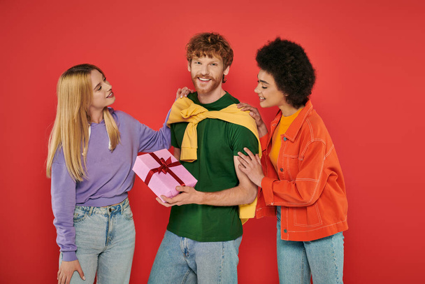 open relationship, happy redhead man holding gift box near interracial bisexual women, polyamorous lovers smiling on coral background, holiday, festive occasions, alternative family  - Photo, Image