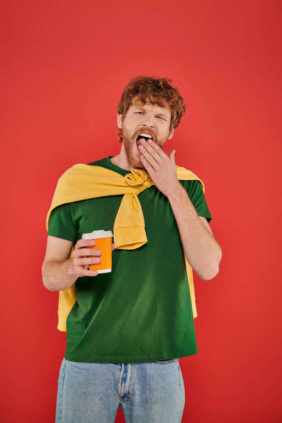 morning coffee, redhead man with beard and curly hair holding paper cup on coral background, vibrant colors, male fashion, takeaway drink, sleepy man yawning and covering mouth  - Photo, Image