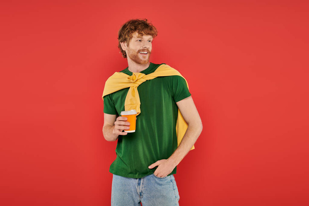 morning coffee, energy, redhead man with beard and curly hair holding paper cup and standing with hand in pocket, on coral background, vibrant colors, male fashion, takeaway drink, hot beverage - Φωτογραφία, εικόνα