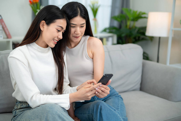 Asian couple sitting on the phone together in the living room at home. Smiling female couple spending quality time together. Lesbian couple love moments happiness concept. - Valokuva, kuva