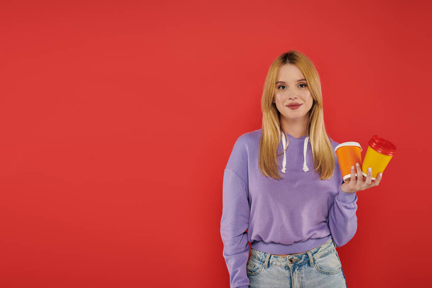 takeaway drink, youthful fashion, blonde young woman in casual attire holding paper cups on coral background, cheerful, looking at camera, vibrant colors, fashion forward, hot beverage  - Foto, Bild