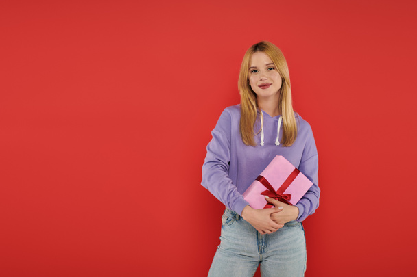 holiday, happiness, blonde young woman in casual attire holding present on coral background, looking at camera, vibrant colors, wrapped gift box, attractive and stylish, festive occasions  - Photo, Image