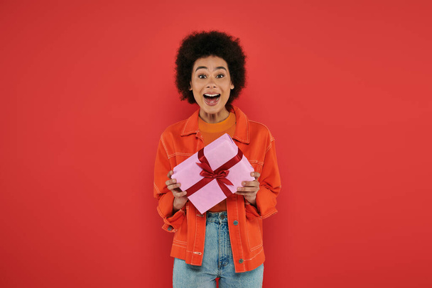 holiday, amazed african american woman in casual attire holding wrapped present on coral background, vibrant colors, gift box, attractive and stylish, festive occasions, looking at camera  - Photo, Image