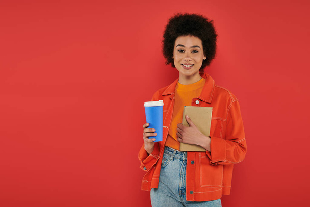 takeaway drink, cheerful african american woman in casual attire holding book on coral background, vibrant colors, attractive and stylish, coffee to go, paper cup, female student looking at camera  - Фото, изображение