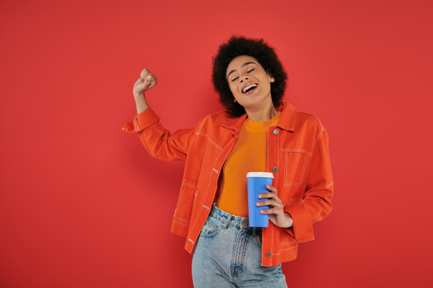 takeaway drink, cheerful african american woman in casual attire holding paper cup on coral background, vibrant colors, attractive and stylish, coffee to go, excited and inspired  - Photo, Image