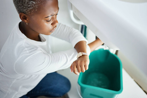 Plumbing, leak and watch with a black woman in the bathroom of her home waiting for repair assistance. Sink, emergency and time with a young female homeowner in her house to stop water using a bucket. - Photo, image